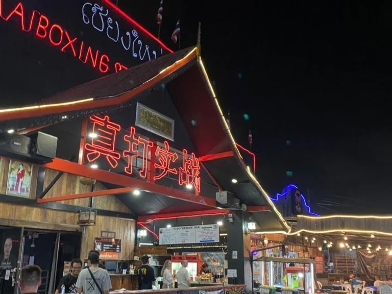 The illuminated entrance of Chiangmai Boxing Stadium at night, featuring neon signage and an inviting atmosphere.