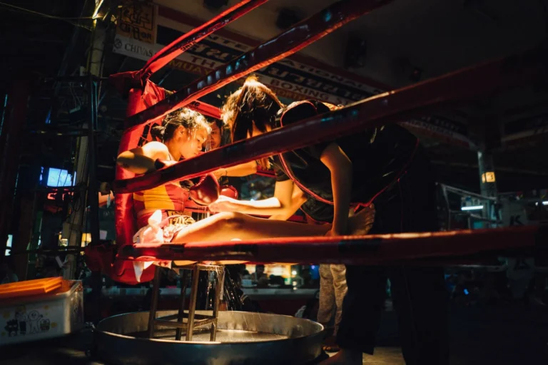 A Muay Thai fighter seated in the ring corner being attended to by trainers during a break, with dramatic lighting emphasizing the scene.
