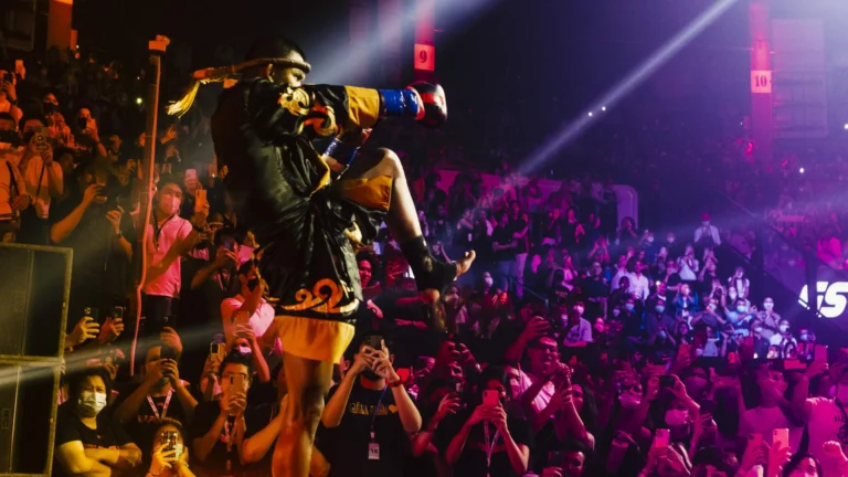 A Muay Thai fighter in black and gold attire performing the traditional Wai Kru dance in front of a cheering crowd.