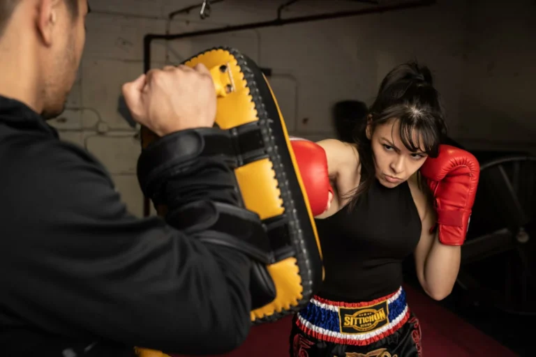 A determined female Muay Thai fighter wearing red gloves and traditional shorts practicing punches with a pad holder in a gym.