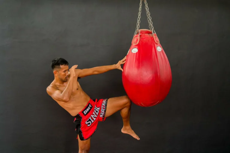 A Muay Thai fighter in red shorts practicing knee strikes with a red heavy bag in a gym setting.