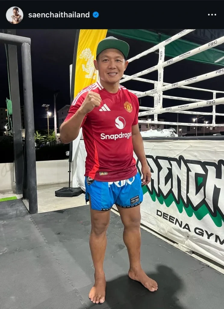 Saenchai, a legendary Muay Thai fighter, posing with a smile near the training ring wearing a red Manchester United jersey and blue shorts.