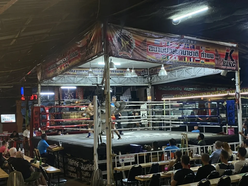 A Muay Thai fight taking place in Thaipae Boxing Stadium at night, with spectators enjoying the match from nearby tables.